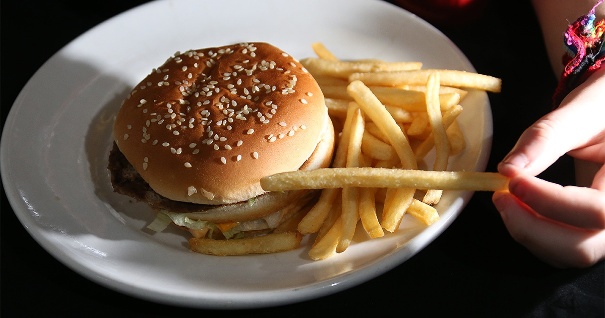 Prato de hambúrguer com batatas fritas, e a mão de uma pessoa pegando uma batata