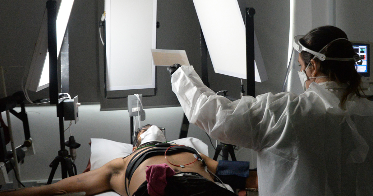 Descrição: pessoa deitada é exposta a três refletores de luz ao seu redor, com medidores no corpo para verificar a pressão arterial e a frequência cardíaca, enquanto uma mulher usando jaleco branco segurando uma ficha supervisiona o teste