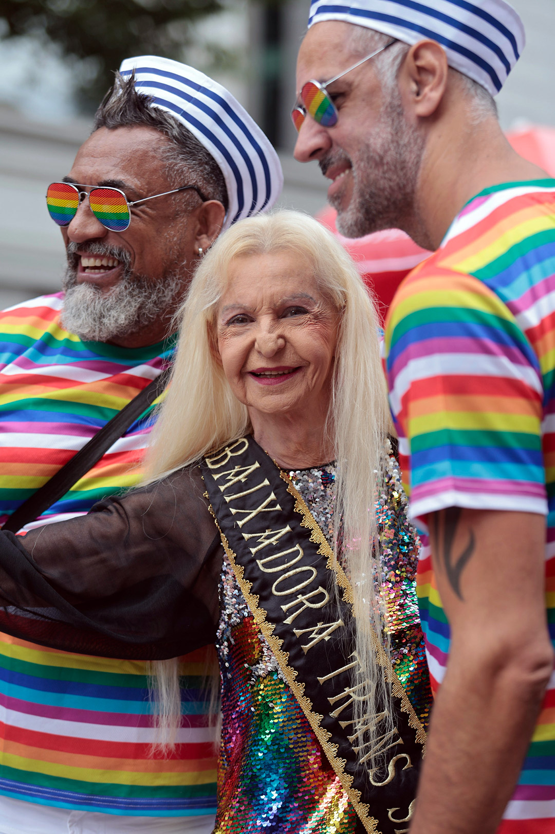 Na Parada LGBT+, dois homens de cabelo e barba grisalhos, com roupas e óculos de arco íris, vestindo um chapéu de marinheiro, estão ao lado de uma mulher idosa com cablos loiros muito longos, que veste uma faixa com os dizeres visíveis "embaixadora trans"
