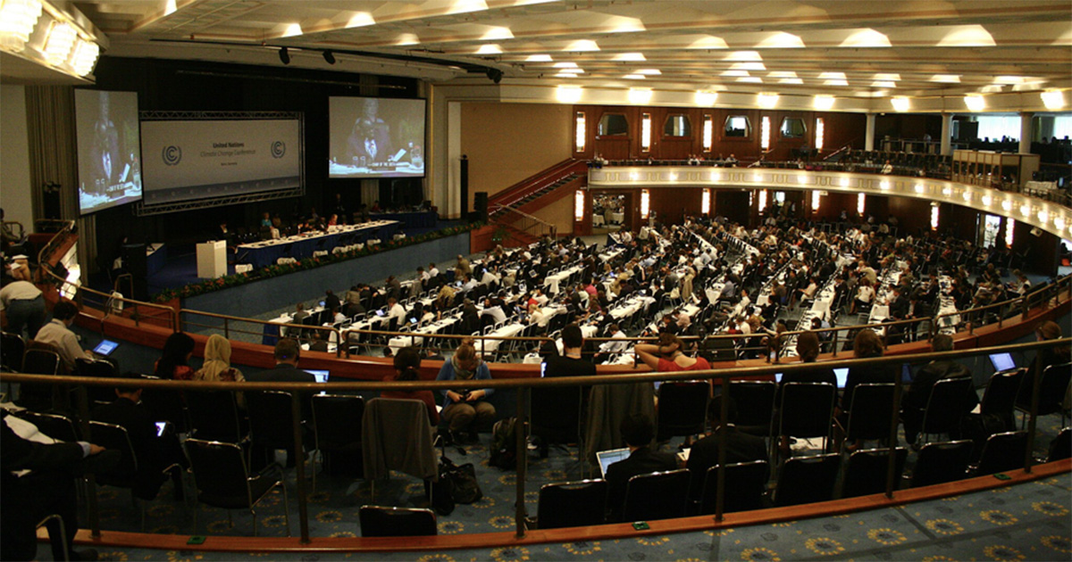 Imagem de uma sala de conferência lotada de pessoas que assistem em telões pronunciamentos de líderes estatais