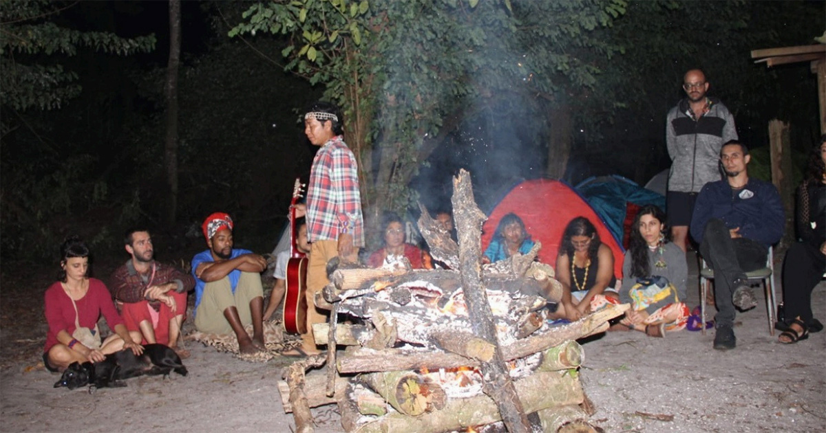 Em uma aldeia, ao redor de uma fogueira, à noite, pessoas variadas se sentam com as cabeças abaixadas. Um home Guarani está levantado ao meio, vestindo uma camisa xadrez e segurando um violão