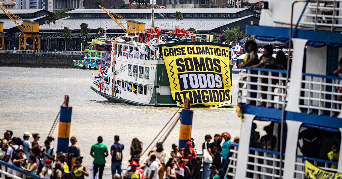 Imagem de uma embarcação em Belém do Pará, durante a COP30, carregando um cartaz com os seguintes dizeres em escritos em preto: Crise Climática Somos Todos Atingidos