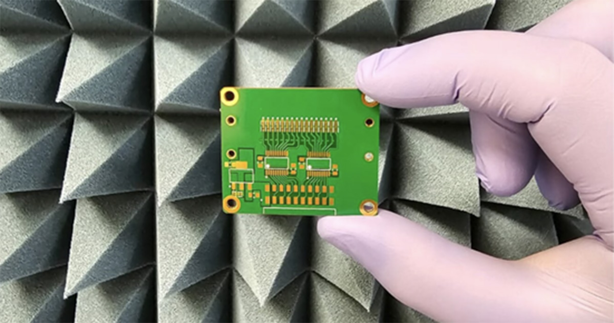 Em frente a uma superfície pontiaguda cinza, mão com luva de cor violeta segura uma placa de circuito impresso usada em dispositivos eletrônicos