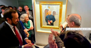 Pessoas em pé aplaudindo e olhando para um quadro na parede com a figura de um homem