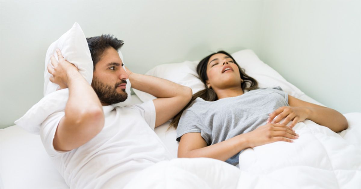 Um casal deitado na cama, em um quarto iluminad. O homem está acordado, visivelmente incomodado, cobrindo os ouvidos com um travesseiro e olhando para a mulher ao lado. A mulher está dormindo de costas, com a boca entreaberta, sugerindo que está roncando. Ambos estão sob um edredom branco, vestindo roupas de dormir.