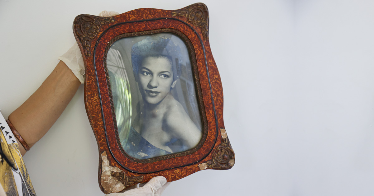 Braços com mãos enluvadas exibem porta-retratos com foto preta-e-branca de uma jovem mulher negra maquiada, usando um vestido tomara-que-caia