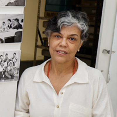 Mulher com cabelo curto e ondulado, com uma mecha grisalha na franja. Usa camisa branca, brincos pequenos e um colar de miçangas vermelhas