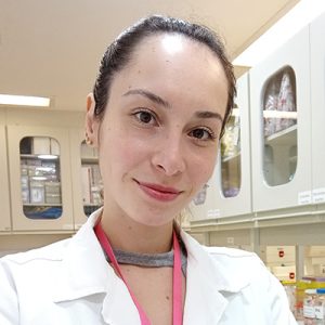 Mulher branca adulta com cabelos escuros presos para trás. Está de braços cruzados e veste um jaleco branco num ambiente de laboratório.