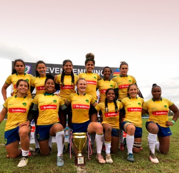 atletas da Seleção Brasileira de Rugby Sevens alinhadas para pose de uma fotografia de time. Estão de pé ou de joelhos sob um gramado e vestem o uniforme da seleção